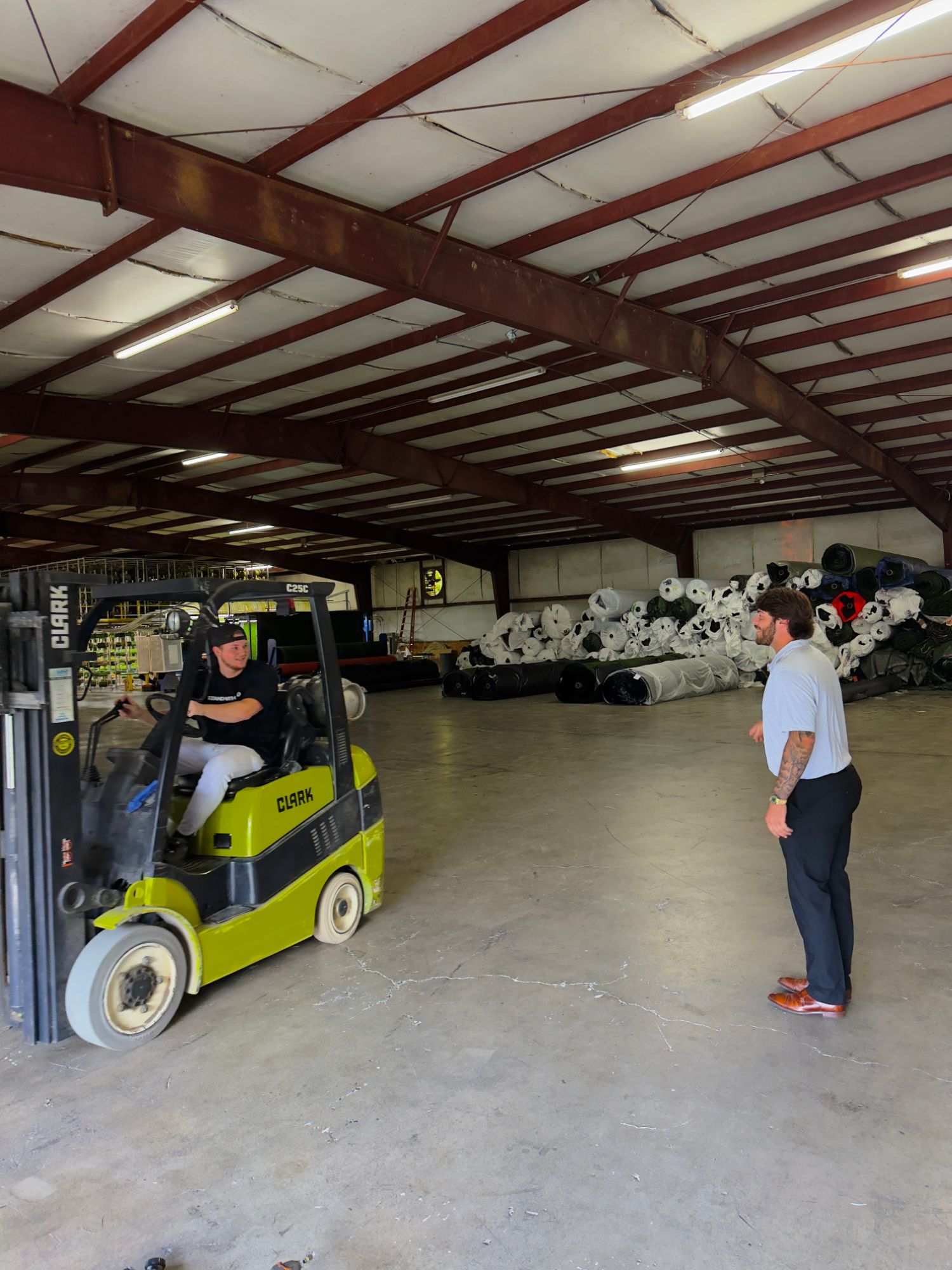Team members talking beside a forklift in the Dalton, Georgia turf warehouse, with wrapped turf rolls stacked behind them