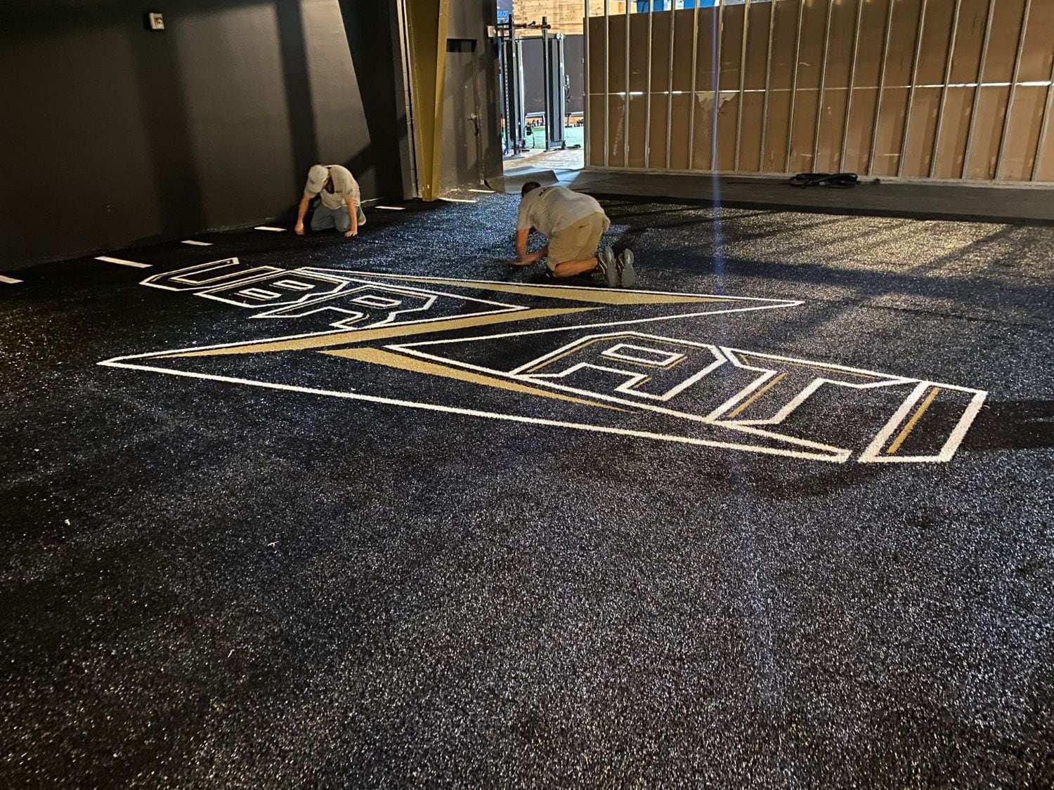 Two installers finishing a black gym turf floor with an inlaid white and gold team logo