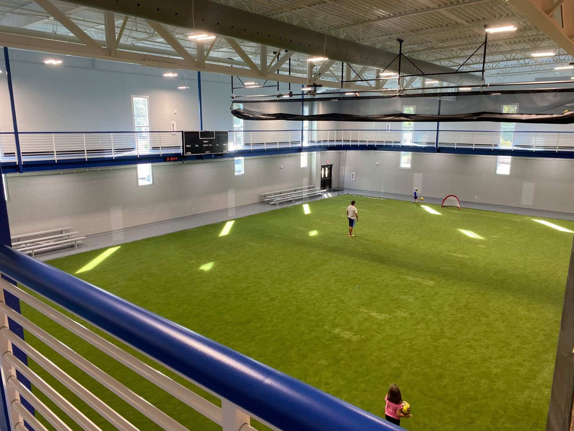 Indoor soccer field on green turf with perimeter netting along the walls, seen from a running-track balcony