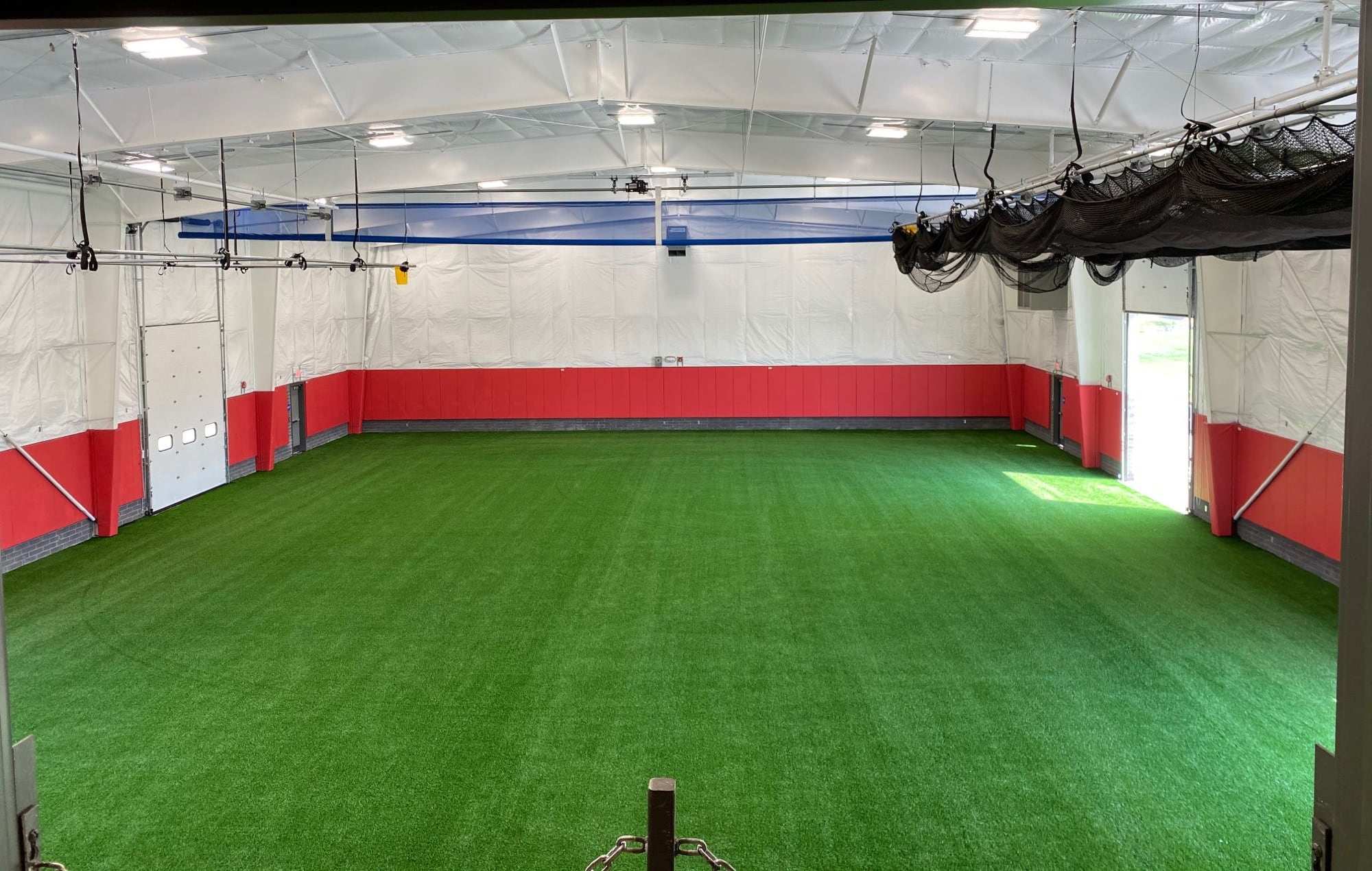 Indoor training facility with green turf, red padded walls and batting cage netting on overhead tracks