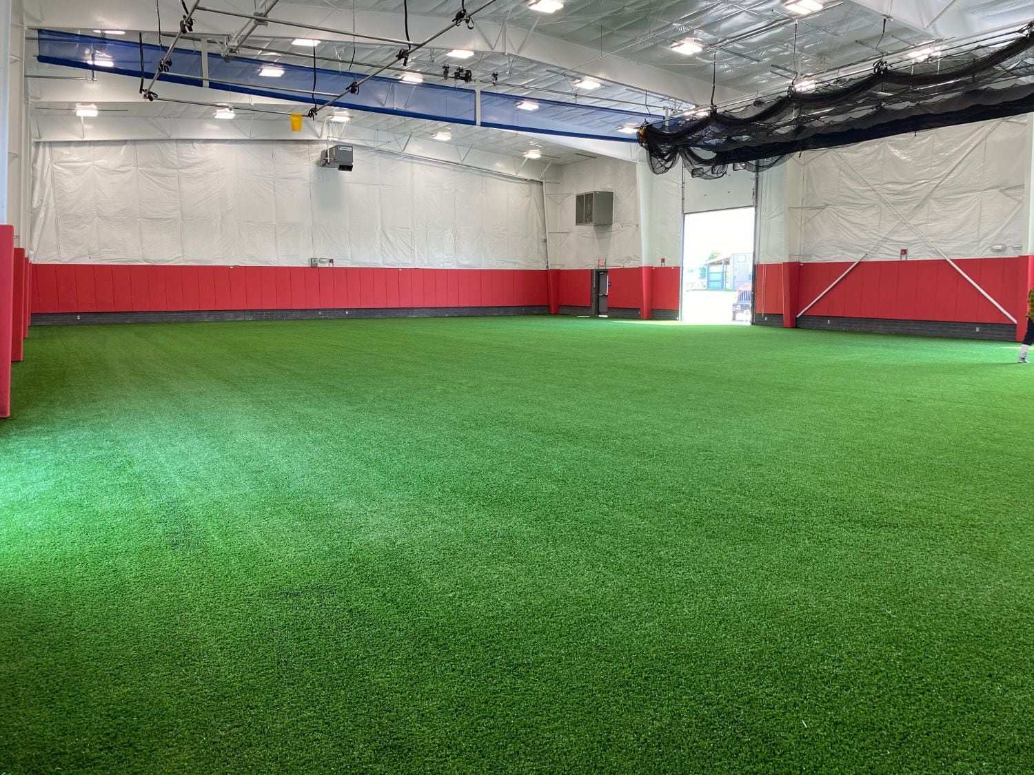 Wide indoor turf floor with red padded walls and a roll-up door to the outdoor fields