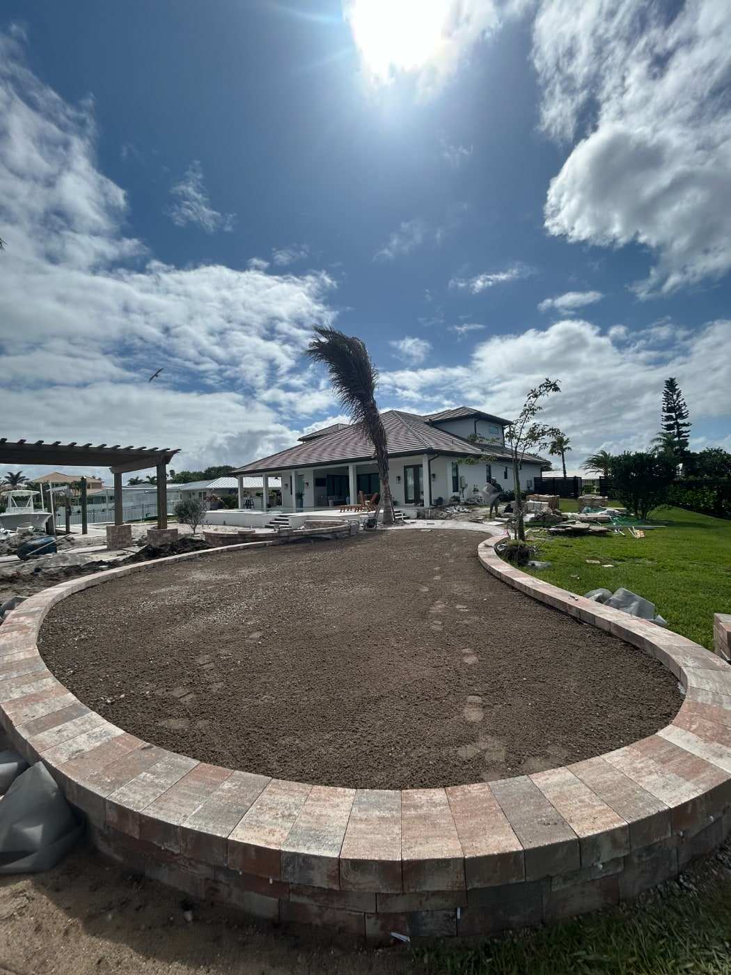 Backyard area excavated down to bare soil inside a curved paver border, with a house and palm tree behind