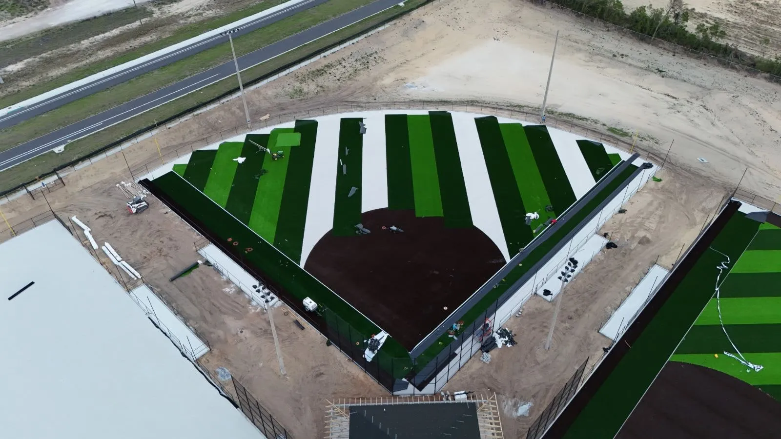 Synthetic turf rolls being laid in stripes across a softball field, with bare base still showing between rows