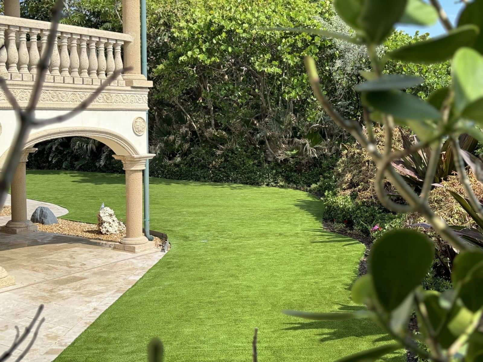 Curved turf lawn beside a stone balustrade and arched loggia, framed by tropical shrubs