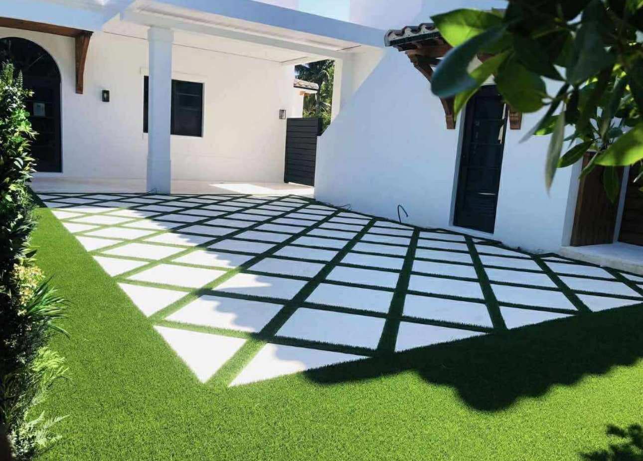 Artificial turf between square white pavers in the front courtyard of a white stucco house