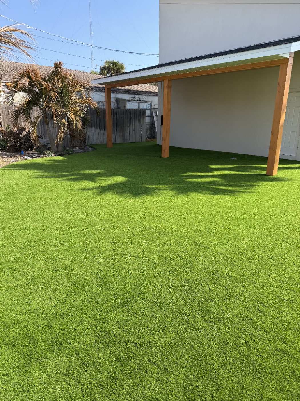 Green artificial turf backyard with a covered patio and a palm tree casting shade across the lawn