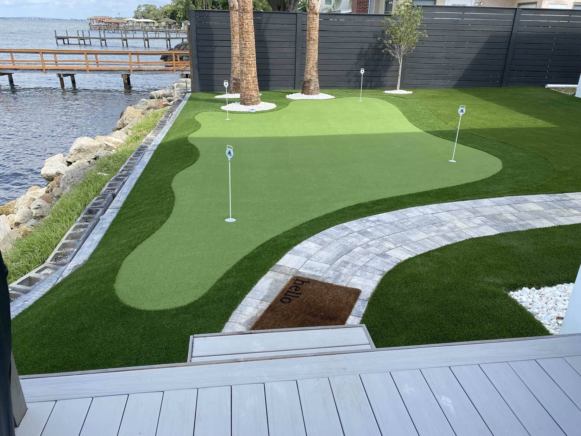 Waterfront backyard putting green with four flagsticks, surrounded by darker landscape turf and a paver walkway beside a dock